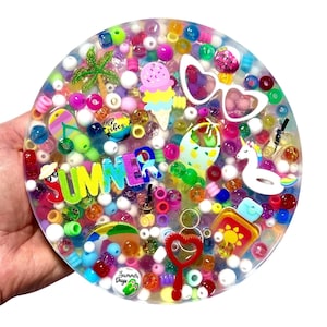 Puede incluir: Un posavasos redondo de resina transparente lleno de cuentas de colores y abalorios con temática veraniega. El posavasos presenta la palabra "SUMMER" en letras brillantes, junto con helados, gafas de sol y un flotador de unicornio. El diseño es vibrante y juguetón.