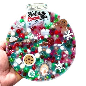 May include: A circular, clear resin ornament with a festive "Holiday Cocoa Bar" design. It features miniature holiday-themed elements such as a gingerbread man, snowflakes, and candy canes, with red, green, and white accents.