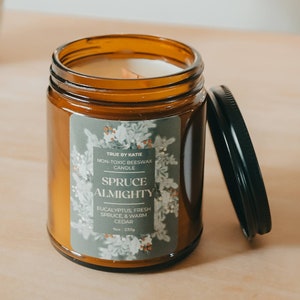 May include: A brown glass candle jar with a white label that reads "TRUE BY KATIE NON-TOXIC BEESWAX CANDLE SPRUCE ALMIGHTY EUCALYPTUS, FRESH SPRUCE & WARM CEDAR". The candle is partially burned and the lid is open.