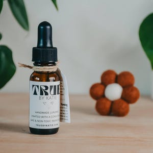 May include: A brown glass bottle with a black dropper and a label that reads "TRUE by Katie". The label also states "HANDMADE LUXURY CRAFTED WITH A CONSCIENCE". A small, flower-shaped felt decoration is to the right.