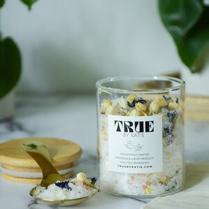 May include: A clear glass jar filled with bath salts and dried flowers, with a gold spoon holding a sample. The jar has a label that reads "TRUE BY KATIE". The lid is made of wood. The background is a marble surface.