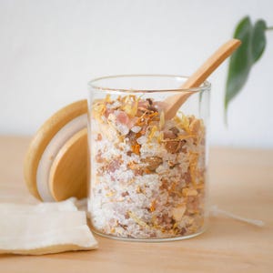 May include: A clear glass jar filled with bath salts, dried flowers, and a wooden spoon. The jar has a wooden lid and sits on a light-colored wooden surface. A small white cloth is next to the jar.