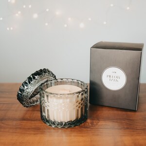 May include: A gray glass candle jar with a lid, filled with a beige candle. The candle jar has a decorative, faceted design. The jar is sitting on a wooden surface. The text on the black box reads "Pillow Talk".