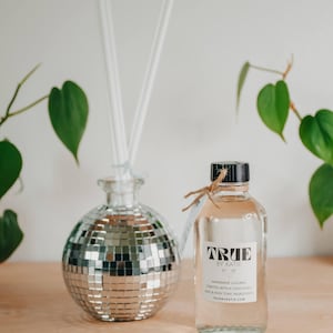 May include: A decorative disco ball-shaped diffuser with white reeds and a clear glass bottle of True by Katie fragrance. The bottle has a black cap and a label with text. Green leaves are in the background.