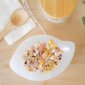 May include: A white, leaf-shaped dish filled with a mix of dried flowers and crystals. A wooden spoon rests on a white cloth, and a wooden lid is visible in the background. The items are arranged on a light wood surface.