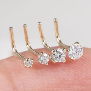 May include: Four L-shaped nose studs crafted from gold-toned metal, each featuring a single round clear gemstone set in a four-prong setting. The studs are arranged in a row, showcasing varying sizes of the stones from smallest to largest.