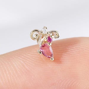 Op de afbeelding: Een kleine, sierlijke oorbel met een goudkleurige metalen basis. Het heeft een peervormige roze edelsteen, een kleine rode edelsteen en een kleine heldere steen. Het ontwerp bevat wervelende, decoratieve elementen.