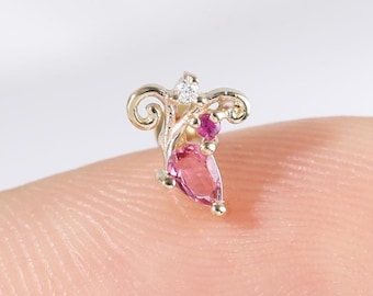 Può includere: Un piccolo orecchino ornato con una base in metallo color oro. Presenta una gemma rosa a forma di pera, una piccola gemma rossa e una minuscola pietra trasparente. Il design include elementi decorativi a spirale.