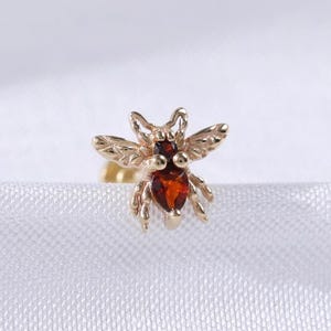 Könnte beinhalten: Ein goldfarbener Ohrstecker in Bienenform mit einem roten Edelstein-Körper. Die Flügel und Details der Biene sind fein gearbeitet. Der Ohrring ist vor einem weißen, strukturierten Hintergrund platziert.