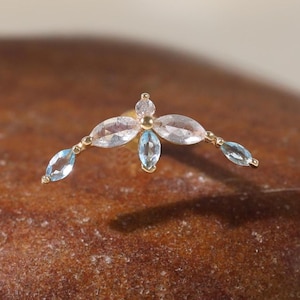 May include: A gold-colored earring with a floral design. The earring features a central clear gemstone surrounded by four light blue gemstones. The earring is set on a brown, textured surface.
