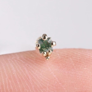 May include: A tiny gold stud earring featuring a green gemstone. The earring has a detailed design with small, round embellishments. The background is a soft, neutral colour, which accentuates the earring's intricate features. The earring is small and delicate.