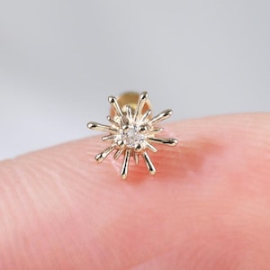 Puede incluir: Un pequeño pendiente dorado con un diseño de estrella. El pendiente presenta una gema central y transparente y puntas radiantes. La joya se muestra en la yema de un dedo, destacando su delicado tamaño y sus intrincados detalles.