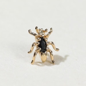 Puede incluir: Un pendiente de botón con forma de insecto en tono dorado que presenta una piedra negra central de talla marquesa incrustada en el cuerpo. La pieza tiene seis patas articuladas y una estructura de cabeza detallada con acabado metálico pulido.