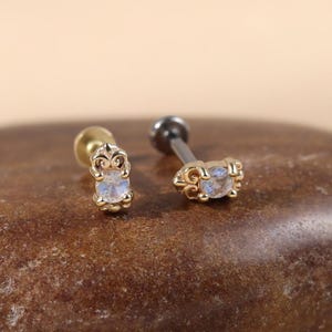 May include: Two gold-toned stud earrings featuring a small, round, iridescent white stone set in a decorative metal frame with intricate scrollwork details. Each earring has a straight post back, showcasing a polished finish and a delicate design.