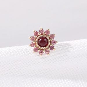 Op de afbeelding: Een goudkleurige oorbel met een bloemmotief. Het midden heeft een donkerrode edelsteen, omringd door een ring van kleinere roze edelstenen. De buitenste bloemblaadjes zijn ook roze edelstenen, waardoor een bloemachtig uiterlijk ontstaat. De oorbel staat tegen een witte achtergrond.