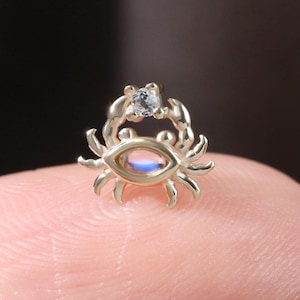 Könnte beinhalten: Ein kleines, goldfarbenes Schmuckstück in Form einer Krabbe mit einem zentralen, oval geformten, irisierenden Stein und einem kleinen, klaren Edelstein oben. Die Krabbe hat detaillierte Beine und Klauen, vor unscharfem Hintergrund.