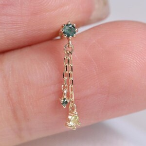 May include: A delicate gold earring with a small, round, teal gemstone at the top, and a chain suspending two smaller gemstones. The lower gemstones are teal and yellow. The earring is designed for a single ear piercing.