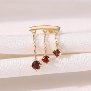 Könnte beinhalten: Ein goldfarbener Ohrring mit drei hängenden roten Edelsteinen. Der Ohrring hat einen horizontalen Balken mit drei Ketten, an denen jeweils ein roter Edelstein hängt. Die Edelsteine sind rund und quadratisch geschliffen. Der Ohrring wird auf weißem Untergrund präsentiert.