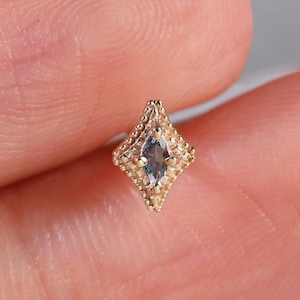 May include: A small gold stud earring featuring a marquise-cut light blue stone set within a diamond-shaped frame. The metal frame has a textured, beaded edge detail surrounding the central stone, creating a geometric, pointed shape.