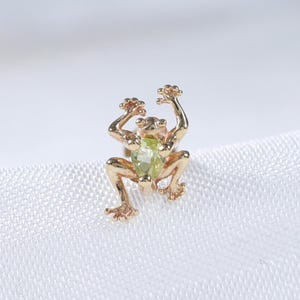 May include: A small gold-toned stud earring shaped like a frog, featuring a light green gemstone set into the center of its body. The frog is depicted in a climbing position with its four limbs extended outward, showcasing detailed texture.