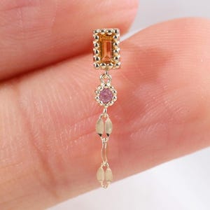 May include: A gold dangle earring featuring a rectangular orange gemstone in a beaded setting, followed by a small round pink gemstone and a delicate gold chain with two flat, elongated gold links hanging vertically.
