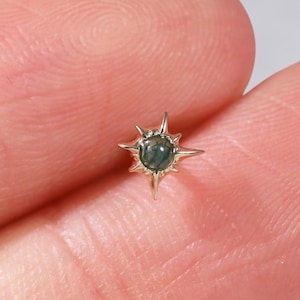 May include: A small gold stud earring featuring a central round dark green gemstone set within a starburst-shaped metal frame with eight pointed rays extending outward from the center, creating a celestial design for a piercing.