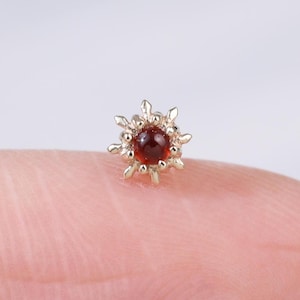 May include: A tiny, gold-coloured stud earring featuring a central, round, dark red gemstone. The earring has a sunburst design around the gemstone. The earring is displayed on a fingertip against a blurred white backdrop.