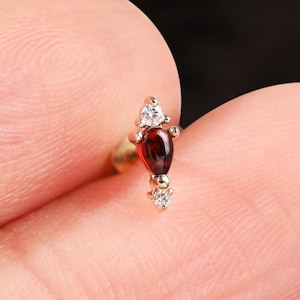 May include: A gold-colored earring with a teardrop-shaped, dark red gemstone at the center, flanked by two smaller, clear gemstones. The earring is held between fingers against a dark background.