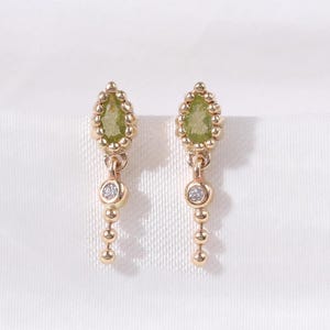 Pode incluir: Um par de brincos pendurados dourados. Cada brinco apresenta uma pedra preciosa verde clara em forma de pêra no topo, um pequeno diamante e uma corrente pendurada de pequenas contas douradas. Em um fundo branco.