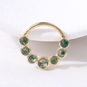 May include: A gold-colored septum ring featuring a series of round, green gemstones. The gemstones are evenly spaced around the ring's circumference. The ring is set against a white background, highlighting the jewelry's design and color.