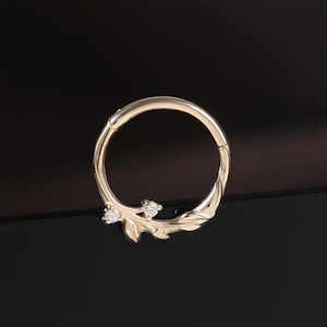 May include: A gold-coloured nose ring featuring a leaf and gemstone design. The ring is circular with a hinged closure. The leaf design has small, clear gemstones. The ring is on a black surface.