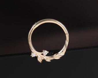 May include: A gold-colored nose ring with a leaf and gemstone design. The ring is circular with a hinged closure. The leaf design has small, clear gemstones. The ring is on a black surface.