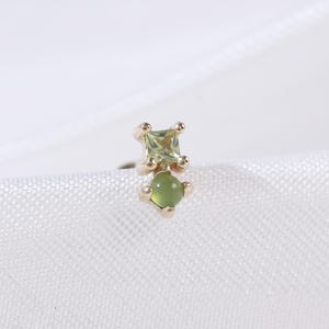May include: A gold-colored earring with two green gemstones. The top gemstone is square-cut, and the bottom is a rounded shape. The earring is set against a white, textured background.