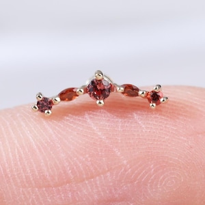 Può includere: Un delicato gioiello curvo con una serie di piccole pietre preziose rosse incastonate in un metallo color oro. Le pietre preziose sono disposte in un arco delicato, con una pietra più grande e rotonda al centro e pietre più piccole su entrambi i lati. Il gioiello è mostrato su un dito.