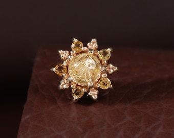 May include: A rose gold stud earring featuring a central round rutilated quartz gemstone surrounded by a halo of smaller, round, yellow-toned gemstones arranged in a starburst pattern with tiny metallic accents between each stone.