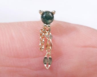 May include: A small, gold-colored earring with a dark green, round stone at the top. The earring features a dangling chain design with small, leaf-shaped charms. The earring is shown on a finger.