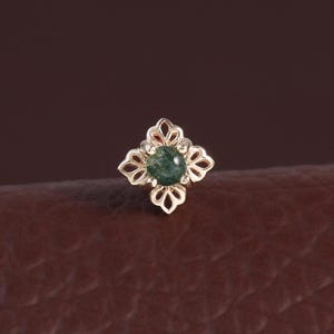 Op de afbeelding: Een kleine, goudkleurige oorbel met een vierbladontwerp. Het midden van de oorbel heeft een ronde, donkergroene edelsteen. De oorbel is geplaatst op een bruin leren oppervlak.