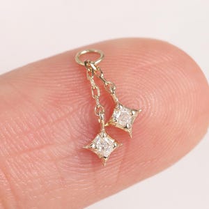 May include: A small, gold-toned earring charm with two star-shaped pendants. Each pendant has a clear gemstone at its centre. The charm is suspended from a small ring. The item is shown against a plain background.