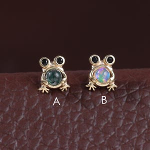 May include: Two small gold frog-shaped earrings. One earring has a green gemstone, and the other has an opal gemstone. The earrings are displayed on a brown leather surface. The earrings are labeled A and B.