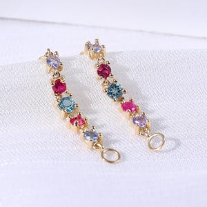 May include: A pair of gold-colored earrings with a linear design. Each earring features a series of colorful gemstones, including red, blue, and purple, set along a slender bar. The earrings are displayed on a white, textured surface.