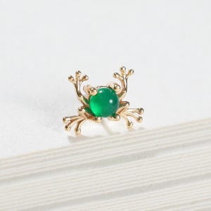 May include: A gold-toned stud earring shaped like a crab with a small, round, vibrant green gemstone set in the center. The metal crab features delicate, thin legs extending outward from the central stone, creating a detailed, miniature design.