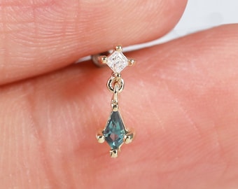 May include: A gold dangle earring featuring a small, clear, square-cut gemstone at the top and a larger, kite-shaped, teal-colored gemstone suspended below. The metal setting is thin and delicate, holding both stones in a secure, pronged arrangement.
