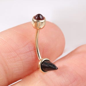 以下が含まれることがあります： ゴールドカラーのへそピアス。カーブしたバーベルデザインで、上部には小さく丸い赤い宝石、下部には黒い円錐形のスパイクが付いています。ジュエリーは柔らかくニュートラルな背景に表示されています。