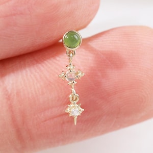 May include: A delicate gold earring with a small, round, green gemstone at the top, and a dangling design of two star-shaped elements, each with a small, clear gemstone. The earring is held between two fingers.
