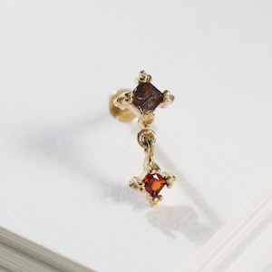 May include: A gold stud earring featuring a dark brown square-cut gemstone at the top and a dangling, smaller square-cut red gemstone at the bottom. The earring has a delicate metal frame with four prongs securing each stone in a vertical arrangement.