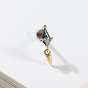 Peut inclure: Une boucle d'oreille à tige en or avec une pierre d'agate mousse en forme de cerf-volant montée sur quatre griffes, accompagnée d'une petite breloque en forme de pointe dorée suspendue. Le bijou présente une finition dorée métallique.