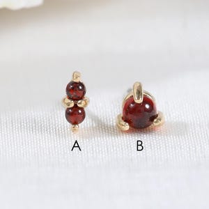 May include: Two gold stud earrings featuring deep red garnet gemstones. Option A displays two small stacked round garnet beads on a vertical gold post. Option B features a single round garnet cabochon set in a gold prong setting. Both are labelled.