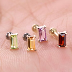 May include: Four stud earrings featuring rectangular baguette-cut gemstones in light green, orange, pink, and deep red. Each earring has a metallic post backing and a prong setting that secures the vibrant, faceted stone in a vertical orientation.