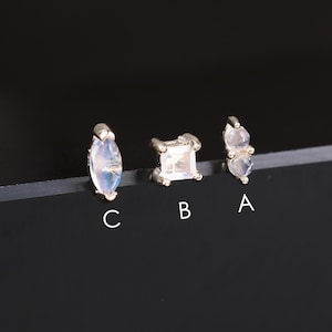 May include: Three small, silver-toned earrings with different gemstone shapes. One earring has a marquise-shaped stone, another a square stone, and the third features two smaller stones. The earrings are displayed on a dark surface.