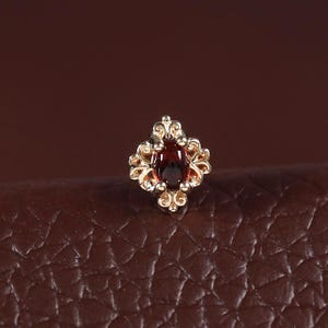 May include: A small stud earring featuring a central oval-shaped deep red gemstone surrounded by a decorative gold-toned metal frame with intricate filigree-like scrollwork details, creating a symmetrical and ornate floral-inspired design.